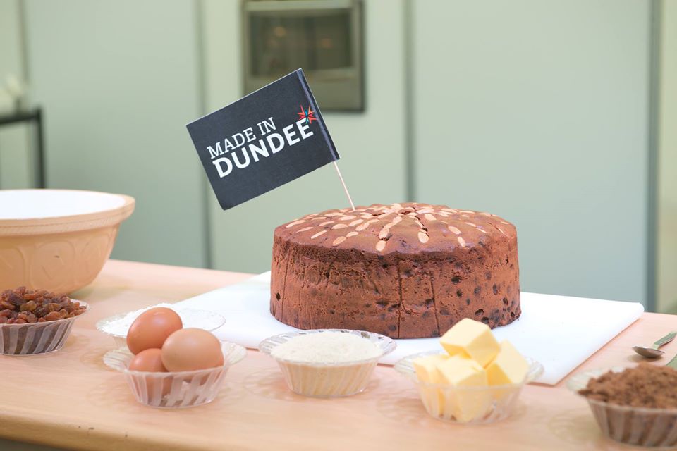 Dundee Cake with flag in the middle of it that is black and says Made In Dundee in white text. Ingredients surround the cake.